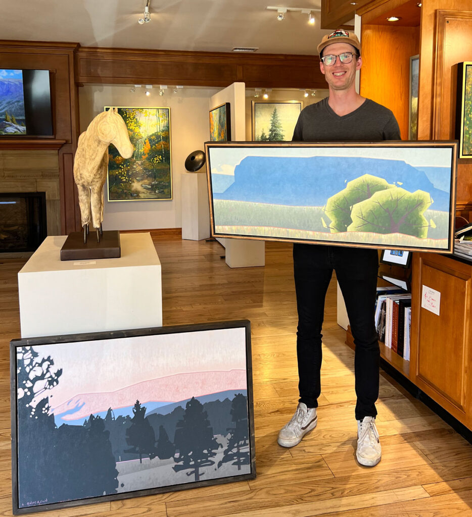 Luke Anderson standing in Ann Korologos Gallery holding a painting of a blue mesa and trees